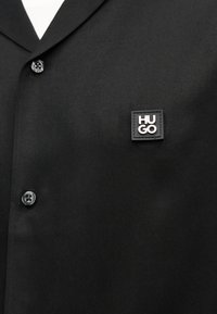 Sort skjorte med knapper og en sort firkantet lap med hvid "HUGO" tekst på venstre brystområde.