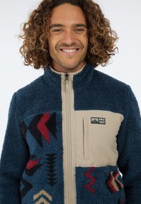 Lächelnder Mann mit lockigem Haar, der eine blaue Fleecejacke mit roten und schwarzen geometrischen Mustern sowie einer beigen Brusttasche trägt, die mit "Protest" beschriftet ist.