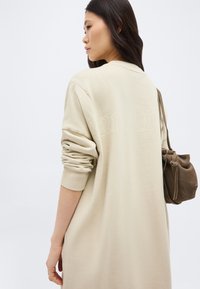 Γυναίκα με μακριά σκουρόχρωμα μαλλιά που φοράει ένα άνετο μπεζ φόρεμα-φούτερ και κρατάει μια καφέ ώμο-τσάντα από σουέτ, στραμμένη προς τα αριστερά με φόντο λευκό.