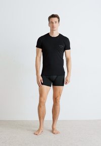 Fekete, rövid ujjú póló, mellkasi logóval, fekete boxer nadrággal párosítva, férfi modellen, egy egyszínű világos háttér előtt.