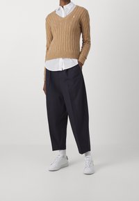 Camelkleurige gebreide trui met kabelpatroon, V-hals en embleem. Witte collared shirt eronder, marineblauwe wijde broek en witte sneakers met gestructureerde zolen.