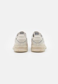 Zapatillas deportivas beige con material texturizado, cuello acolchado y suela de goma distintiva con patrones de agarre. Construcción robusta con detalles de ventilación.