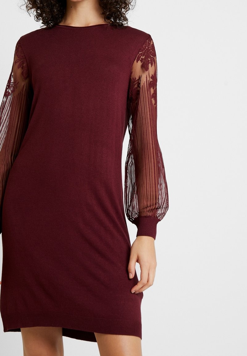 Donna che indossa un abito borgogna con lunghe maniche in pizzo trasparente, decorate con ricami floreali e polsini a costine.