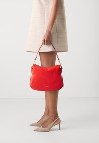Borsa a spalla in pelle rossa con superficie texturizzata, hardware dorato e una maniglia singola, tenuta da una persona in un vestito a quadri chiari e tacchi nudi.