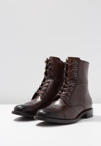 Botines de cuero marrón hasta el tobillo con punta redonda, cordones en la parte delantera, costuras sutiles y tacón bajo tipo bloque. Textura suave y diseño clásico.