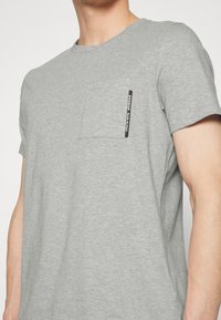 Homme portant un t-shirt à manches courtes gris clair avec une petite poche sur la poitrine et une étiquette noire indiquant "DONDUP MADE IN ITALY."