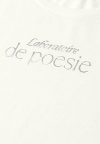 Biała tkanina z błyszczącym srebrnym napisem „Laboratoire de poésie” wydrukowanym blisko kołnierzyka delikatną, kursywą czcionką.