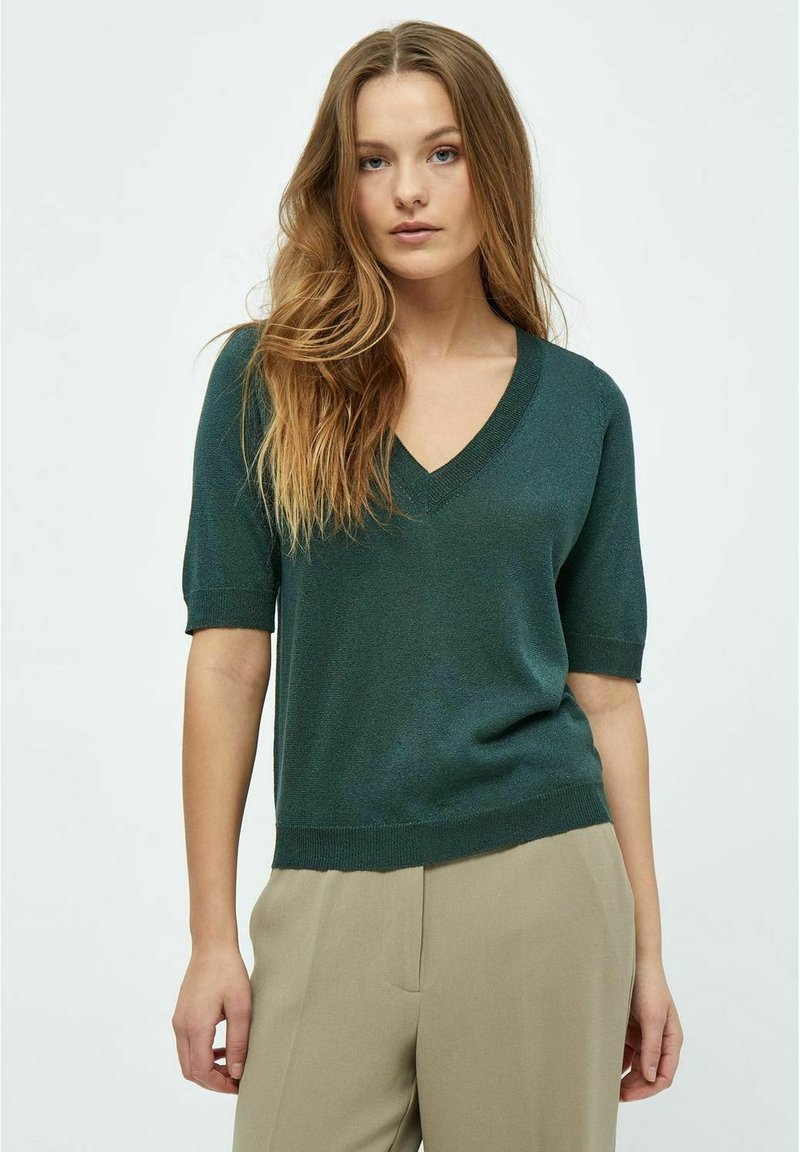 Suéter verde de manga corta y escote en V en una tela ligera y texturizada, combinado con pantalones beige neutros, creando una silueta relajada.