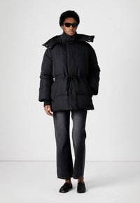 Black puffer jacket with a belted waist, high collar, and detachable hood. The model wears dark jeans and black loafers.