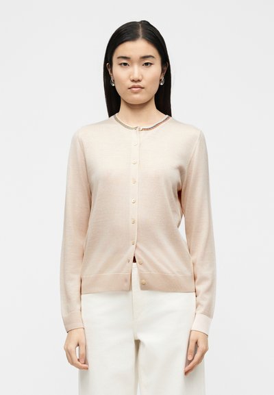 Γυναίκα που φοράει ένα ανοιχτό ροζ ζακέτα με κουμπιά και πολύχρωμη μπορντούρα στον γιακά, καθώς και λευκό παντελόνι, που στέκεται μπροστά σε απλό λευκό φόντο.