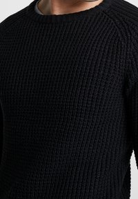 Czarny sweter z dzianiny o teksturowanym, prążkowanym wzorze. Posiada okrągły dekolt i raglanowe rękawy, ukazując gruby, miękki materiał.