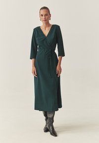 Robe verte à manches trois-quarts, avec une ceinture à la taille et une jupe longue, associée à des bottes noires hauteur genou.