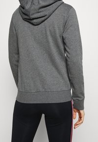 Sweat à capuche gris avec une poche kangourou, une capuche à cordon ajustable et des poignets côtelés. Associé à des leggings noirs avec des bandes latérales roses.