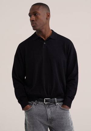 Homme portant un polo noir à manches longues et un jean gris, mains dans les poches, regardant vers la gauche sur un fond uni.