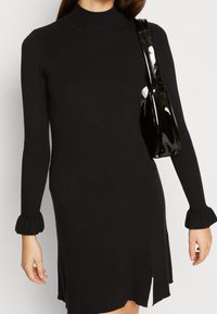 Musta ribbineulottu mekko, jossa on mallikaulus, pitkät hihat ja liehutusresorit hihoissa; sivuhalkio, ja mukana kiiltävä musta laukku.