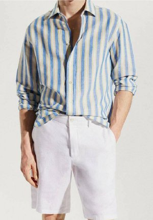Homme portant une chemise à rayures verticales bleues, beiges et blanches avec manches retroussées et un short blanc, les mains partiellement dans les poches.