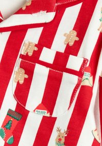 Punavalkoraidallinen kangas, jossa on piparkakkutalokuvioita, sukkia, poroja ja pieni etiketti. Pehmeä puuvillamateriaali, näkyvissä taskut.