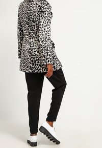 Persona vistiendo una chaqueta de estampado de leopardo en blanco y negro, pantalones negros y zapatos de plataforma blancos, de pie de perfil con una pierna ligeramente levantada.