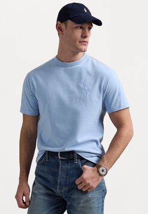 Light blue short-sleeve t-shirt with a subtle logo, paired with faded blue jeans and a dark cap. Features a brown watch on wrist.