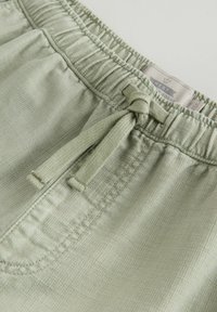 Pantalon à taille élastique vert clair avec cordon de serrage noué et couture de poche avant visible sur un tissu texturé.