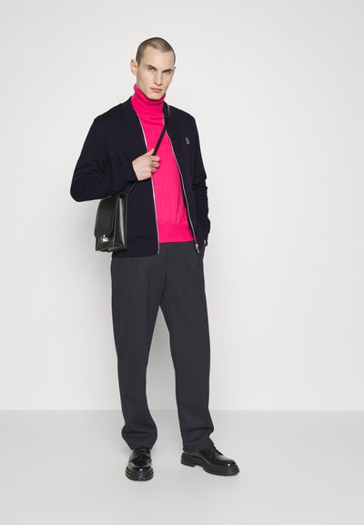 Vīriešu apģērbs, kas ietver tumši zilu zip-up jaku, spilgti rozā turtleneck, ogļu krāsas bikses un melnas kurpes, papildināts ar melnu somu.