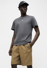 Άνδρας που φοράει κοντομάνικη μπλούζα σκούρου γκρι, μαύρη καπέλο και μπεζ σορτς, στέκεται με το ένα χέρι στην τσέπη, κοιτάζοντας προς το πλάι.