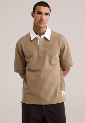 Jeune homme portant un polo beige à manches courtes avec un col blanc, debout les mains derrière le dos devant un fond uni.