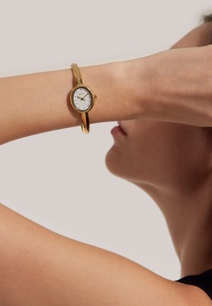 Guld armbåndsur med en cirkulær hvid urskive, minimale markeringer og et slankt design. Remmen er glat og skinnende og sidder tæt på håndleddet.
