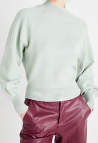 Personne portant un pull col montant côtelé vert pâle et un pantalon en cuir bordeaux taille haute, les mains dans les poches.