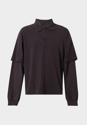 Homme debout portant une chemise noire longue à manches longues, superposée et oversized, un pantalon noir ample et des chaussures en cuir noires sur un fond clair uni.