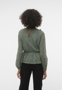 Blusa verde chiaro con un intricato motivo in pizzo, maniche lunghe e dettaglio a oblò sulla schiena. Vita elasticizzata per un look aderente.