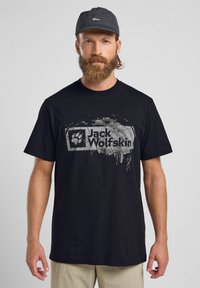 T-shirt nera in cotone con scollo rotondo, caratterizzata da una stampa grafica grigia con "Jack Wolfskin" e logo della zampa sul fronte. Stile semplice.