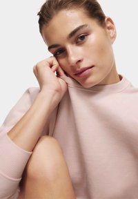 Lichtroze sweatshirt met een ronde halslijn en geribbelde manchetten, met een zachte textuur. Model leunt met de kin op de hand, huid is natuurlijk.