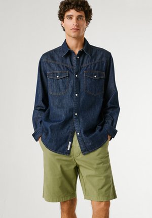 Joven con cabello rizado que lleva una camisa de mezclilla azul oscuro y pantalones cortos de color verde claro hasta la rodilla, de pie con las manos en los bolsillos.