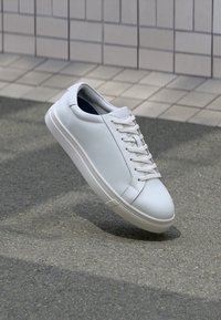 Weißes Leder-Sneaker mit Schnürsenkeln schwebt über einem strukturierten Asphalt vor einem gefliesten Wandhintergrund.