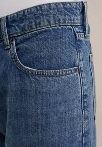 Blauwe spijkerbroek met een klassieke snit, subtiele textuur en oranje stiksels. Inclusief een achterzak met een koperen klinknagelaccent.