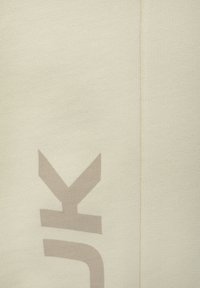 Lichtbeige stof met een gestructureerd oppervlak. Opvallend groot lichtbruin logo gedrukt op de voorkant. Verticale naad die langs de zijkant loopt.