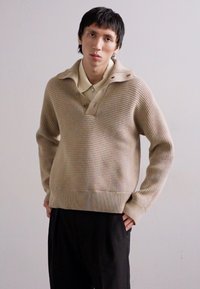Beige ribbed knit sweater with a notch collar, featuring a textured finish and contrasting horizontal stripes. Paired with black pants.