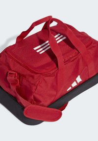 Bolsa de deporte roja hecha de tela duradera, con logotipo blanco y detalles de franjas, base negra, asas dobles y un bolsillo con cremallera.