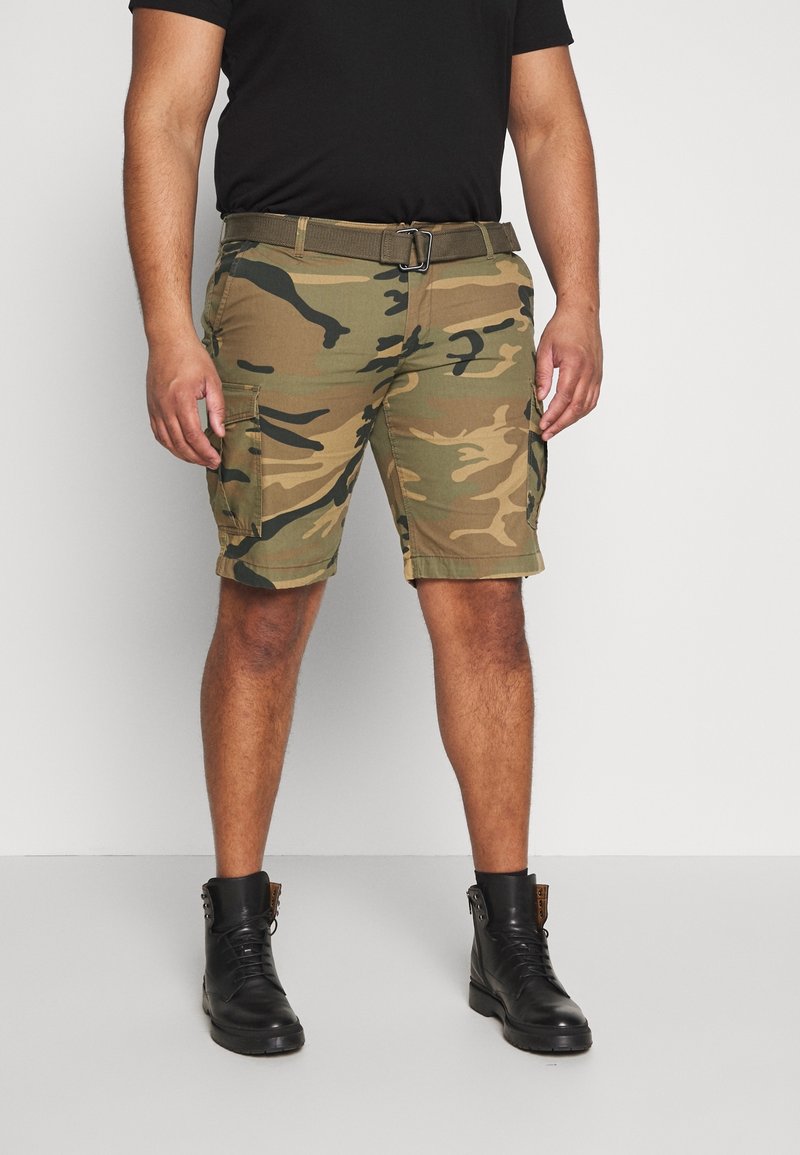 Homme portant un short cargo camouflage, un t-shirt noir, une ceinture marron et des bottes noires à lacets, debout devant un fond uni.