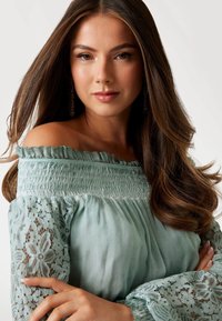 Eine Frau trägt ein hellgrünes schulterfreies Kleid mit Rüschen. Die Ärmel sind mit aufwendigen floralen Spitzenmustern verziert, die Textur hinzufügen.