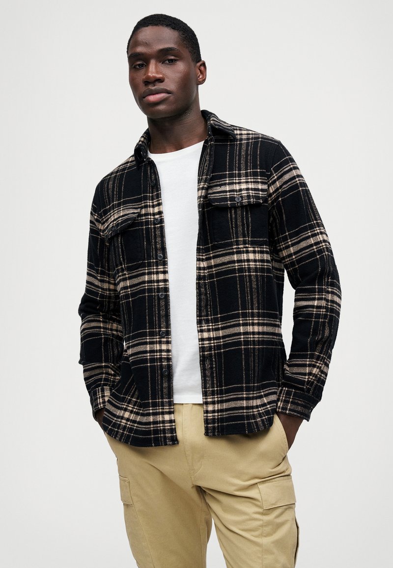 Camisa de franela a cuadros en negro y beige con dos bolsillos en el pecho, cierre de botones en la parte delantera y corte relajado, llevada encima de una camiseta blanca y pantalones beige.