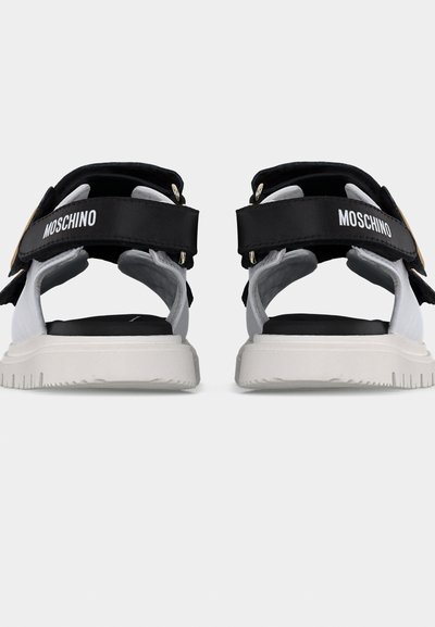 Ζευγάρι ασπρόμαυρων σανδαλιών Moschino με ρυθμιζόμενες λουριά και λευκές σόλες, εμφανιζόμενα από πίσω σε απλό φόντο.