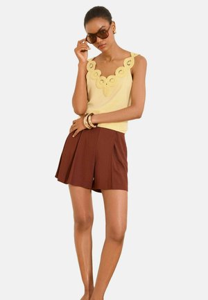 Nő barna pliszírozott rövidnadrágot és sárga ujjatlan felsőt visel hullámos csipkés nyakkivágással, nagy barna napszemüveget igazít.