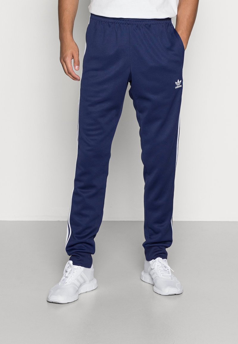 Tmavě modré sportovní kalhoty s bílými pruhy po stranách, vyrobené z hladké látky. Mají elastický pás a elegantní, zúžený střih.