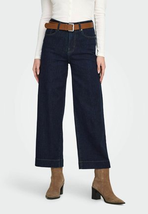 Femme portant un jean large taille haute bleu foncé, ceinture en cuir marron, bottines beiges en daim à talons et haut blanc ajusté boutonné.