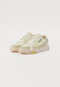 Beige und weiße Sneaker mit limonengrünen Akzenten, durch perforierte Bereiche und einer klobigen Sohle. Mit Schnürverschluss und Branding auf der Zunge.