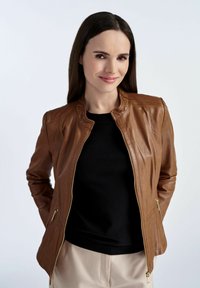 Brązowa skórzana kurtka o dopasowanym kroju, z zamkiem z przodu i kieszeniami po bokach, w zestawieniu z czarną bluzką i kremowymi spodniami.