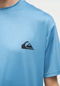 Ανοιχτό μπλε κοντομάνικη μπλούζα με ένα μικρό μαύρο λογότυπο κύματος και βουνού στο αριστερό στήθος, φορεμένη από ένα άτομο μπροστά σε απλό φόντο.