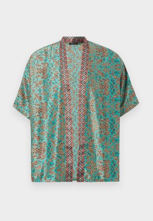Homme portant une chemise ouverte à motif turquoise sur un t-shirt bleu marine, jean foncé, mocassins noirs et portant un sac d'épaule noir.